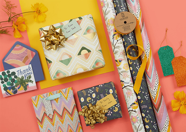 A collection of wrapped presents, rolls of gift wrap and greetings cards, laid flat on a pink and yellow background. 