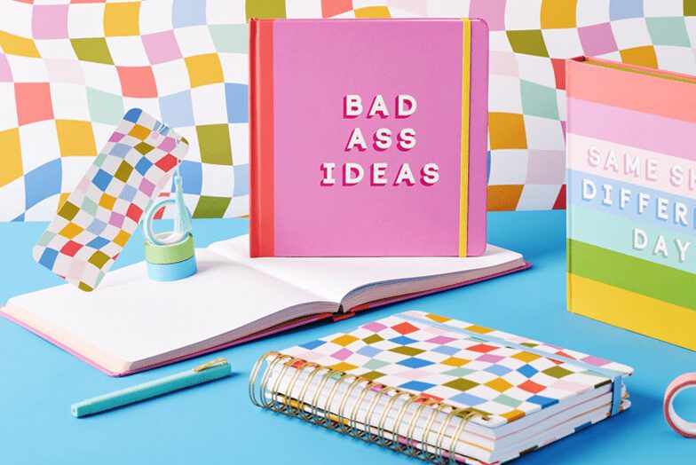 A range of notebooks and pads, on a blue surface with a multicoloured background. The featured book reads "Bad Ass Ideas".
