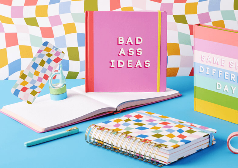 A range of notebooks and pads, on a blue surface with a multicoloured background. The featured book reads "Bad Ass Ideas". 