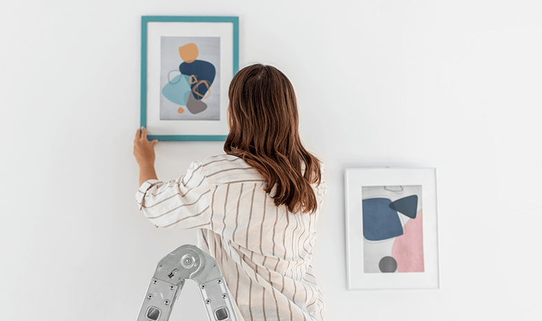 A lady stands on a step ladder and hangs a photo frame on a white wall. 